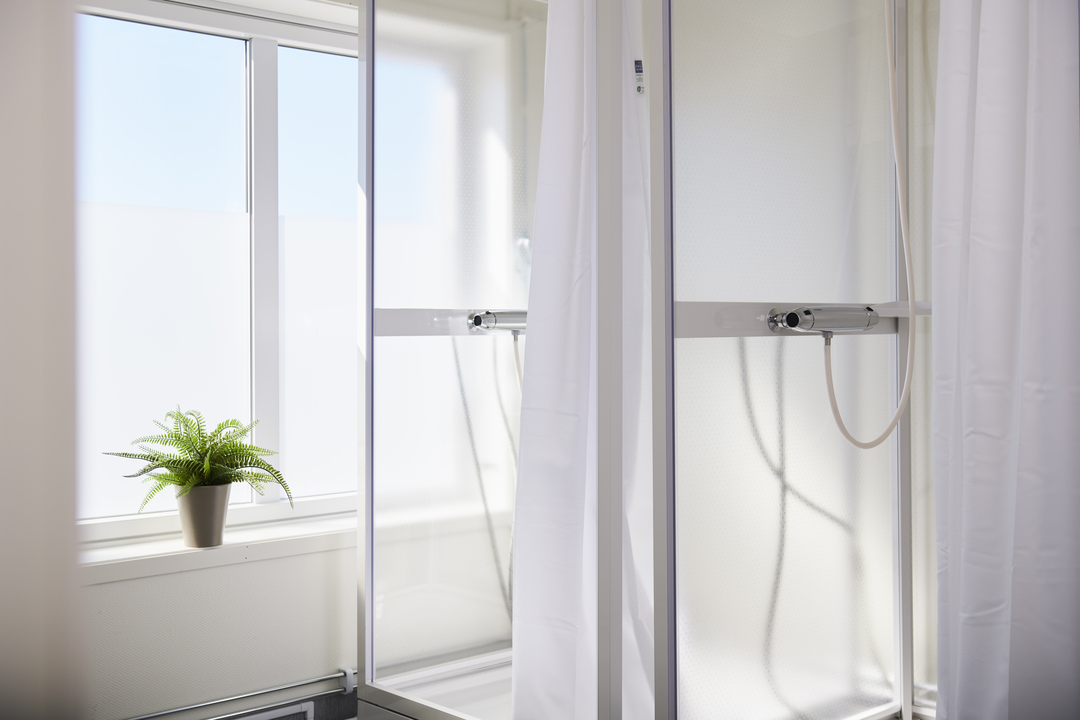 Duschutrymme i anslutning till omklädningsrum i kontorsmoduler från Expandia. Ljusa duschväggar med draperi, handdusch och ett fönster som släpper in dagsljus. Bilden är tagen i kontorsmoduler vid Alviks trafik i Luleå.