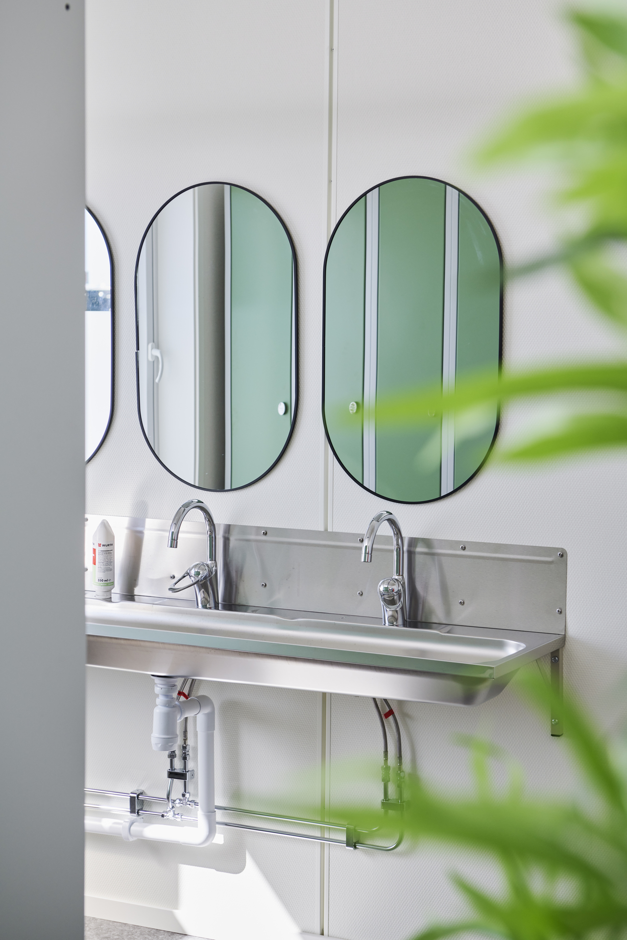 Tvättställ med två handfat och speglar i ett omklädningsrum i kontorsmoduler från Expandia. Ljusa ytor och gröna skåp speglas i speglarna, med växter som mjukar upp miljön. Bilden är tagen i kontorsmoduler vid Alviks trafik i Luleå.