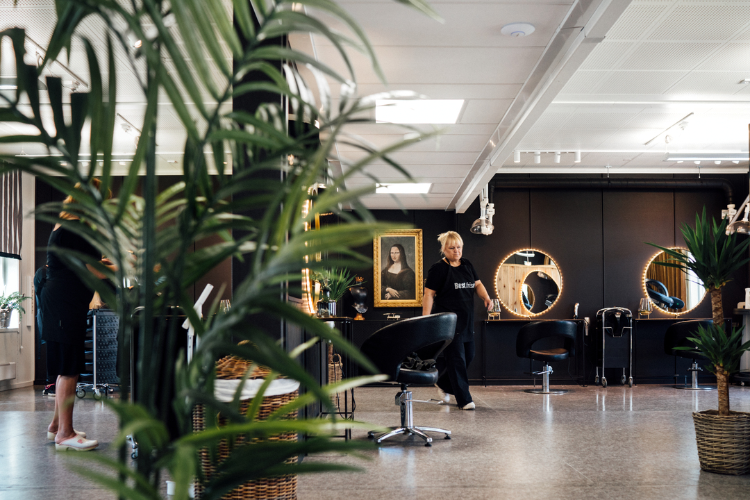 En ljus och modern frisörsalong i Slemmestad centrum i Norge, med flera stylingstolar och runda speglar med belysning längs en mörk vägg. En frisör rör sig genom lokalen, och stora gröna krukväxter står i förgrunden och runtom i rummet. Inredningen är stilren med öppna ytor, och i bakgrunden syns arbetsstationer och salongens utrustning.