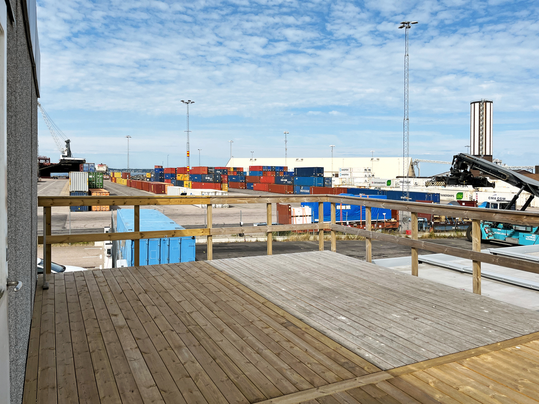 Träaltan med räcke utanför modulerna, med utsikt över ett hamnområde fyllt av staplade containrar, lyftkranar och industribyggnader under blå himmel med tunna moln.
