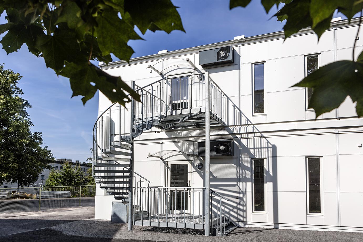 Exteriör av Sveaplansskolan i Stockholm, uppförd med skolmoduler från Expandia. Tvåvånings vit modullösning med raka fasader, höga smala fönster och externa spiraltrappor i stål. Modulerna är placerade i en grön stadsmiljö och visar Expandias flexibla och funktionella modulkoncept för skolor.
