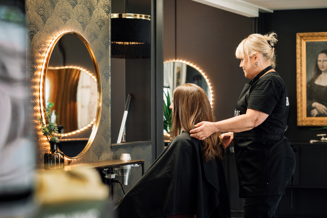 En frisör står bakom en sittande kund i en stilfull salong och förbereder håret inför klippning eller styling. Kunden bär en svart frisörkappa och sitter framför en rund spegel med inbyggd belysning. Miljön är mörk och elegant med dekorativa detaljer, en arbetsbänk med hårverktyg och en tavla på väggen i bakgrunden. I Slemmestad centrum i Norge.