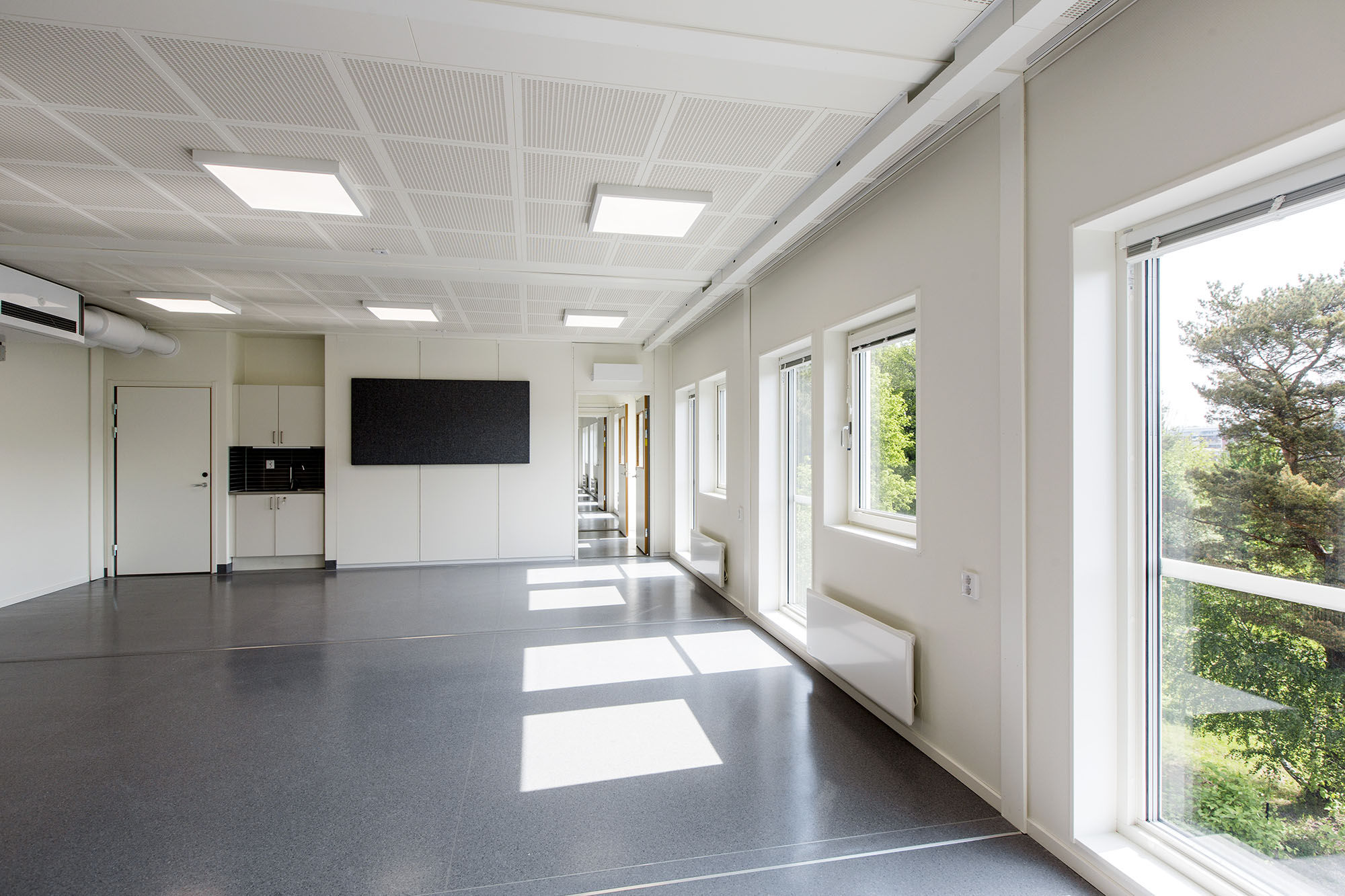 Ljust och öppet klassrum i skolans modullösning med stora fönster längs ena väggen, ljusgrått golv, akustiktak, förvaringsskåp och genomgång till korridor i bakgrunden.