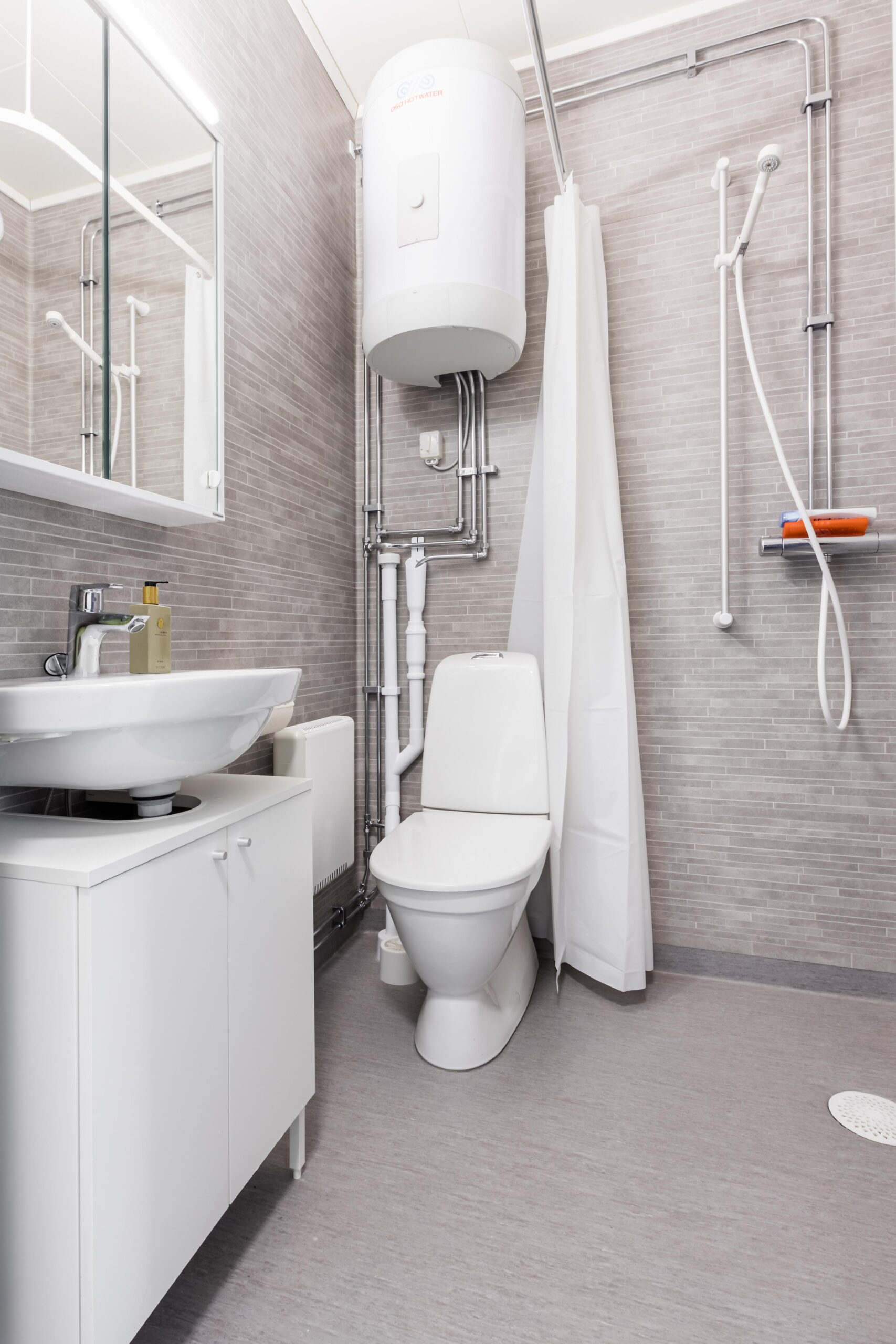 Ljust badrum med toalett, handfat, dusch och varmvattenberedare i korridorsboende.