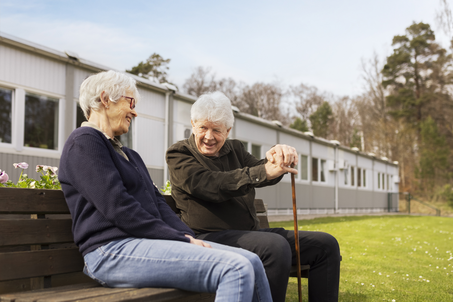 Två boende sitter på en bänk i utemiljön vid Borggården demensvård i Olofström. I bakgrunden syns en modullösning från Expandia som omger gräsytan.