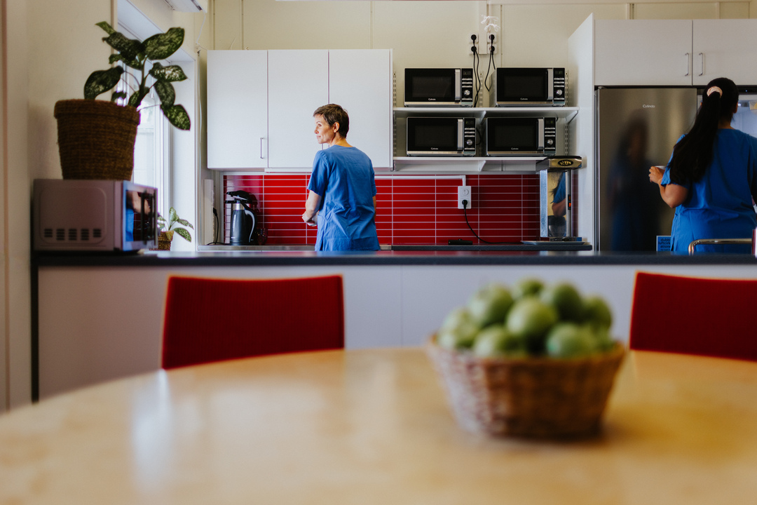 Omsorgspersonal arbetar i ett gemensamt kök vid Herrhagsgården vård- och omsorgsboende i Falun. Köket har ljusa skåp, röda kakelplattor och utrustning för gemensamma måltider, med ett matbord i förgrunden.