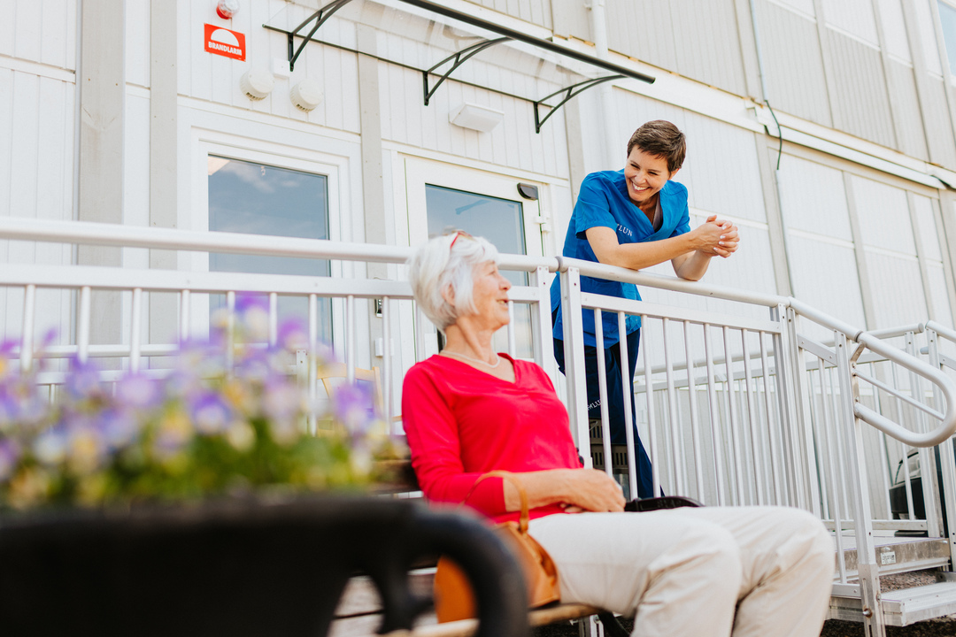 En person sitter utomhus vid Herrhagsgården vård- och omsorgsboende i Falun och samtalar med omsorgspersonal som står vid ett räcke. I förgrunden syns blommande växter och i bakgrunden moduler som ingår i boendets modullösning.