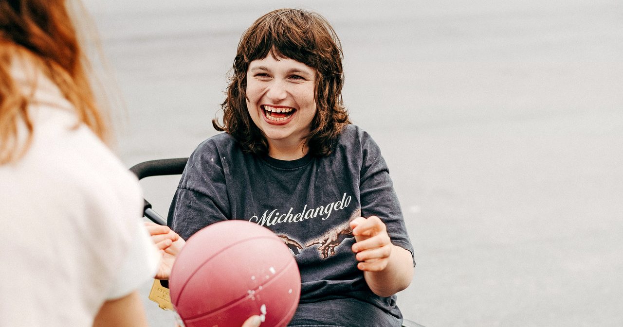 En person i rullstol skrattar och sträcker sig efter en röd boll som räcks fram av en annan person under en gemensam aktivitet utomhus.