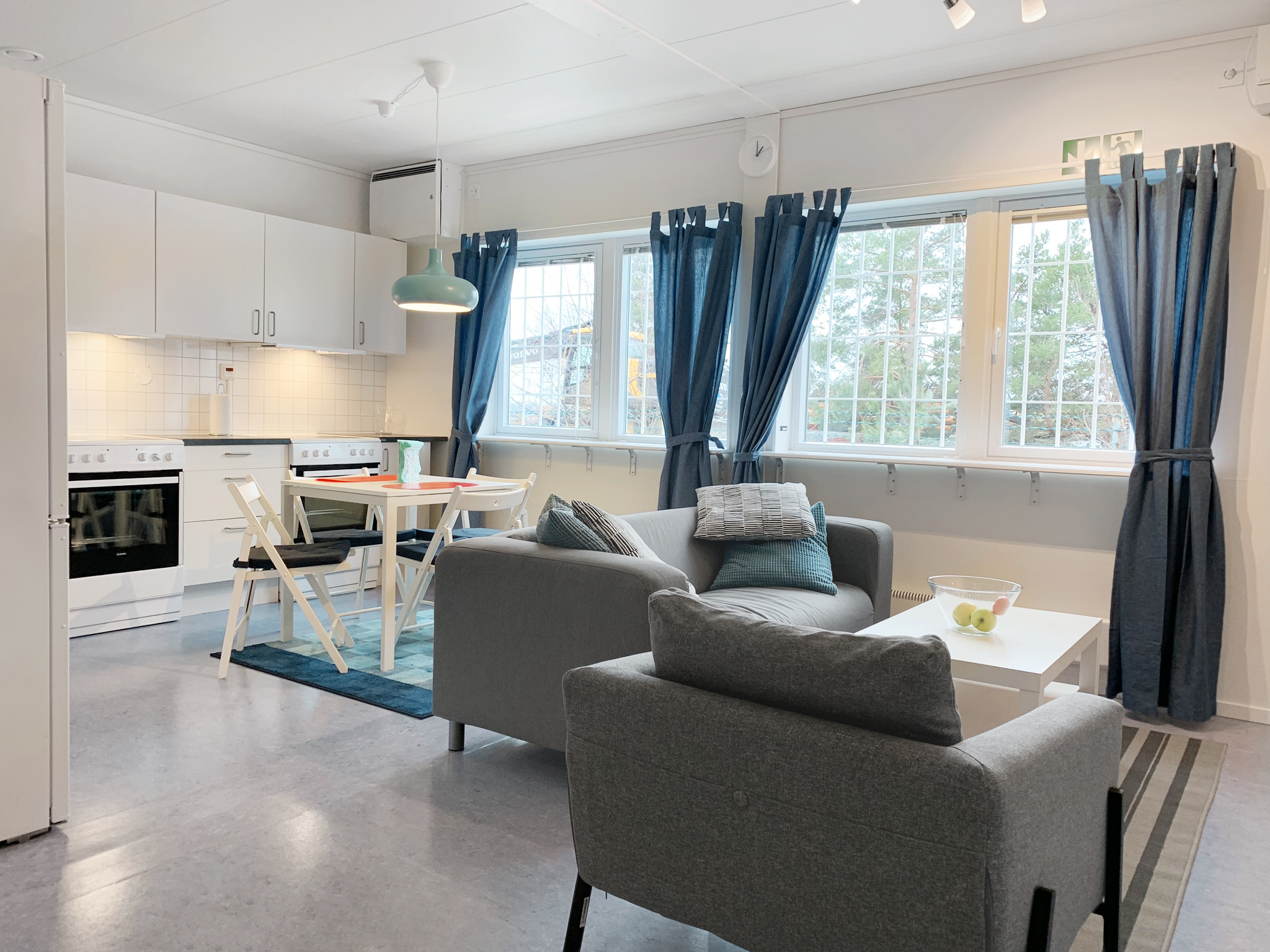 Interiör av ersättningsboende med öppen planlösning, kombinerat kök och vardagsrum, matplats för fyra, soffgrupp, stora fönster med gardiner och ljus, modern inredning i neutrala färger.