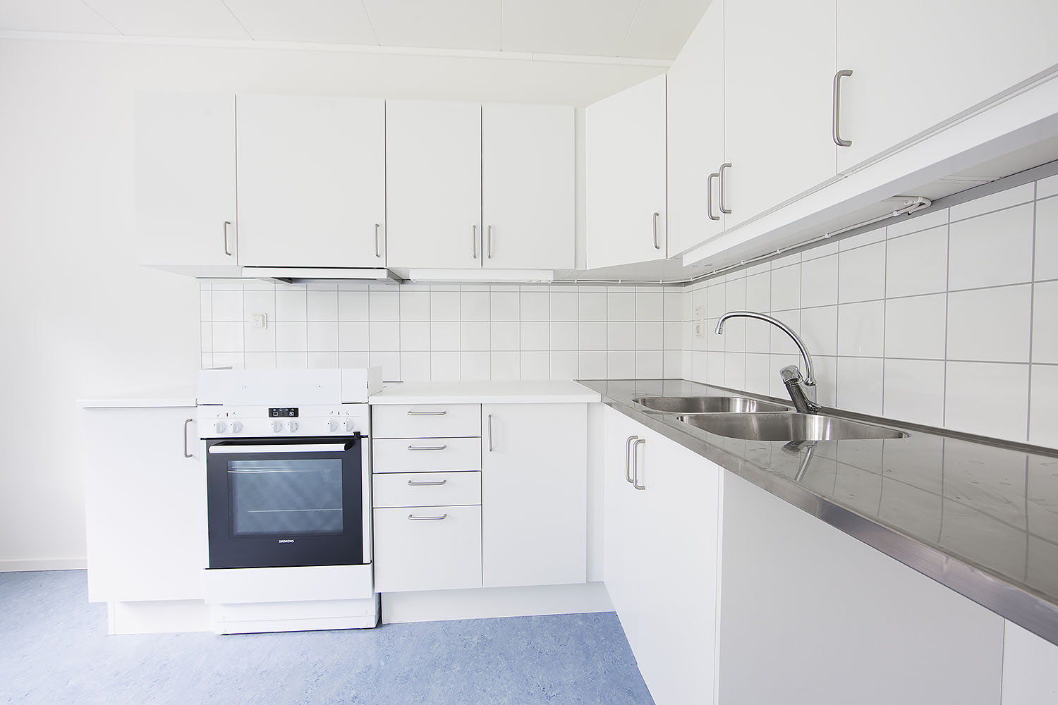 Ljust kök i boendemodul från Expandia med vita skåp, spis med ugn, rostfri bänkskiva och dubbla diskhoar.